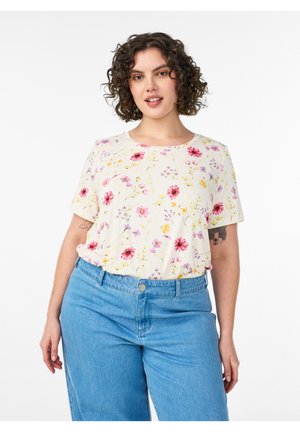 Kvinde med krøllet hår iført en cremet blomstret kortærmet top indstukket i højtaljede blå jeans, stående mod en hvid baggrund.