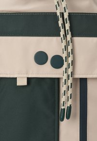 Gros plan sur la poche avant d'un sac à dos beige et vert foncé avec deux boutons-pression sombres et deux cordons à motifs beige et vert.