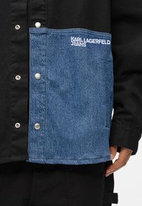 Melnā jaka ar denima kabatu. Kreisajā pusē ir pogas ar spraudņiem un uz denima baltā krāsā izšūts "KARL LAGERFELD JEANS".