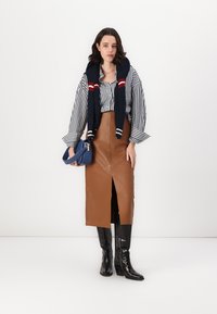 Gonna midi in finta pelle marrone con spacco frontale, abbinata a una camicia a righe bianco e nera e a un maglione blu navy indossato sulle spalle.