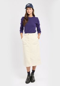 Cremfarbene Cord-Midi-Rock mit zwei aufgesetzten Vorder-Taschen, kombiniert mit einem dunkelpurpurfarbenen Pullover und schwarzen Knöchelstiefeln. Blaue Kappe mit Logo.