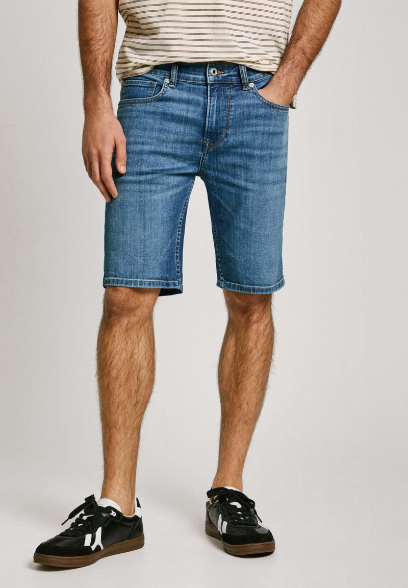 Shorts de mezclilla en azul medio con corte recto, que cuentan con cinco bolsillos y un desvanecimiento mínimo. Combinados con zapatillas negras con detalles blancos.