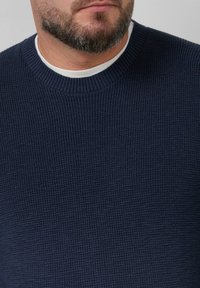 Granatowy sweter dziany o prążkowanej teksturze i okrągłym dekolcie, nałożony na białą koszulę. Zbliżenie na detale tkaniny.