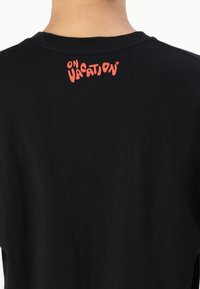 Black cotton shirt featuring a crew neckline and a logo in red reading "ON VACATION" printed on the back near the neck.