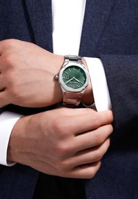 Reloj de acero inoxidable con una esfera texturizada verde, agujas plateadas y acentos en oro rosa, montado en una pulsera de metal plateado.