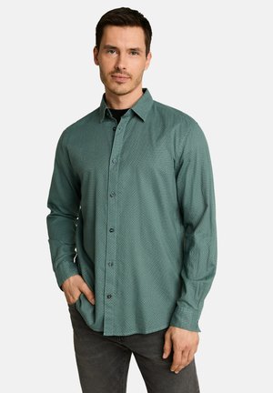 Homme portant une chemise verte à motifs à manches longues boutonnée et un pantalon foncé, posant avec une main dans la poche devant un fond uni.