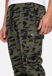 Grønne og sorte camo-joggers, lavet af blødt stof, med lynlåslommer og en cargo-lomme med et logo-patch.