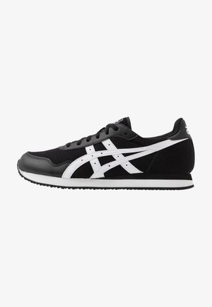 Zapatilla deportiva baja en negro y blanco con rayas blancas en forma de cruz, cordones negros y suela texturizada.