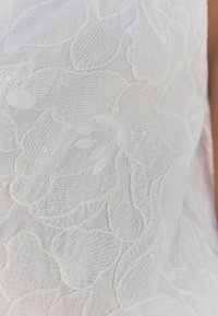 Tissu blanc brodé avec un motif floral aux pétales texturés et des accents subtils de perles. Texture douce et légère dans l'ensemble.