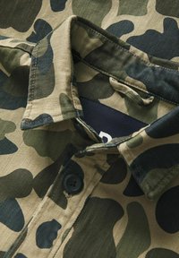 Camisetta a fumetto con colletto, in tonalità verde scuro e beige, con chiusura a bottoni e tessuto textures. Dettagli di cucitura visibili.