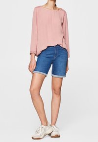 Hellrosa langärmliges Oberteil mit lockerem Schnitt und rundem Ausschnitt, kombiniert mit blauen Jeansshorts mit umgeschlagenen Säumen. Weiße Turnschuhe vervollständigen das Outfit.