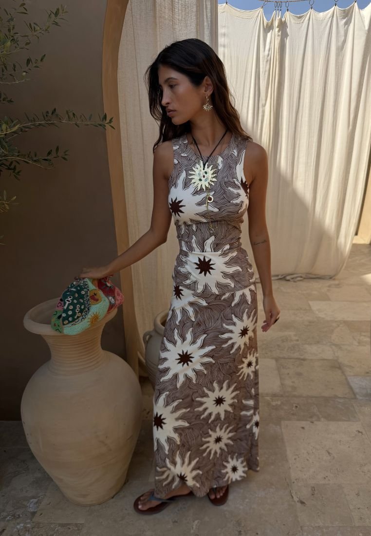 Γυναίκα με αμάνικο μακρύ καφέ και λευκό φόρεμα με σχέδιο ήλιο στέκεται ξυπόλητη σε πέτρινο πάτωμα, στηρίζοντας το χέρι της σε μια μεγάλη μπεζ κεραμική βάζα.