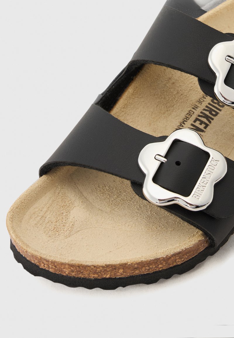 Sandale noire avec deux larges straps, ornée de boucles en forme de fleur en argent. Le lit de pied est en liège beige, avec une surface texturée pour le confort.