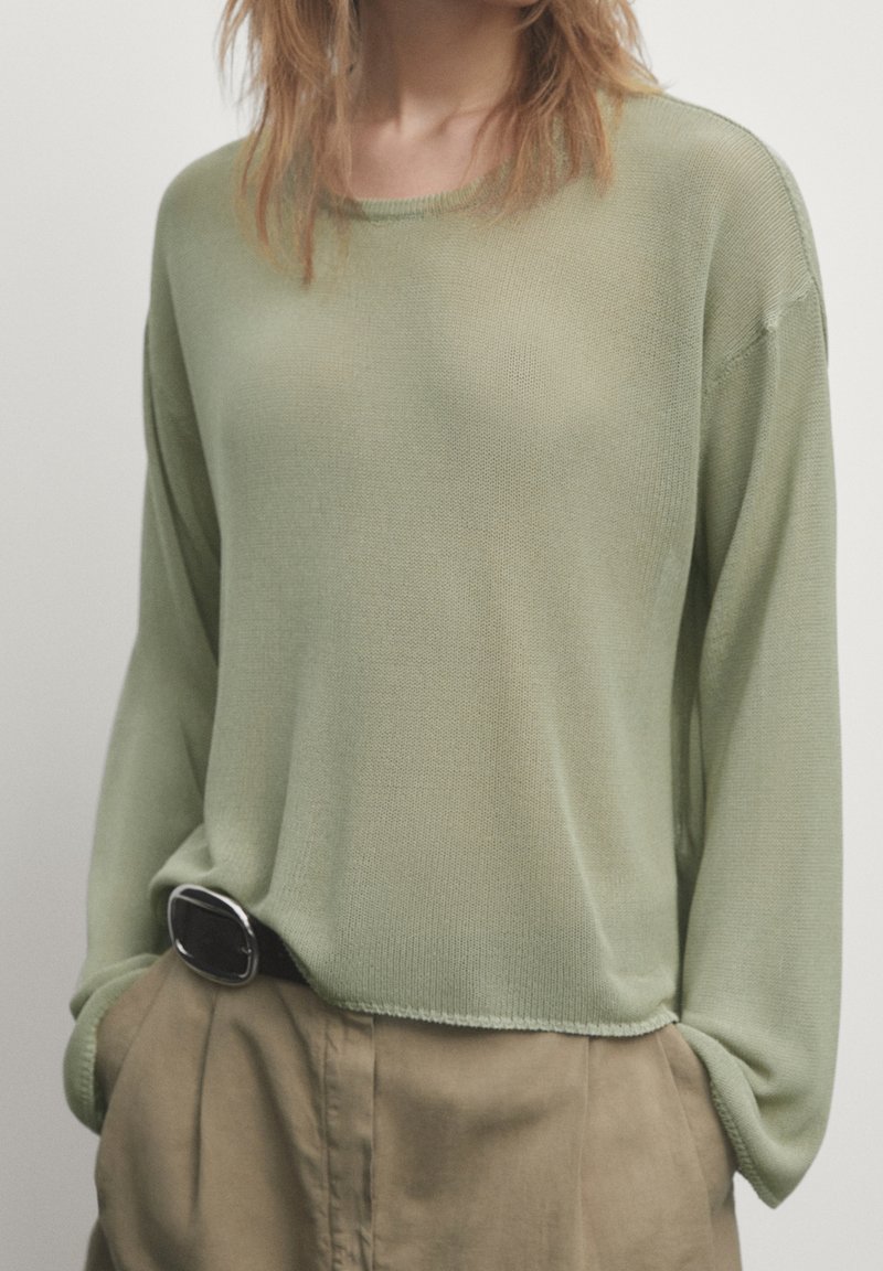 Haut en maille à manches longues de couleur vert clair avec une coupe ample et un tissu texturé. Il présente un col rond et des épaules tombantes, associé à un pantalon beige.