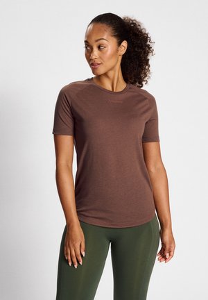 Kvinde med krøllet hår i hestehale iført brun kortærmet træningsbluse og grønne leggings, kigger til venstre mod en hvid baggrund.