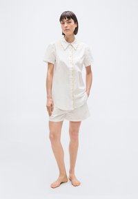 Femme debout pieds nus, portant une chemise blanche à manches courtes boutonnée et un short assorti avec un bord festonné jaune sur le col et à l'avant.