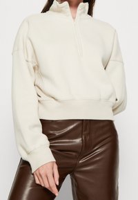 Sudadera de forro polar color crema con cuello alto, cierre frontal de medio zip y ajuste holgado. Cuenta con puños y cintura acanalados, combinada con pantalones de cuero marrón.