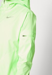 Kurtka Nike w jasnym neonowym zielonym kolorze, z teksturowanym wykończeniem, pełnym kapturem i bezpieczną kieszenią na zamek na lewym ramieniu.