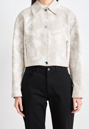 Kobieta w krótkiej beżowej kurtce w kwiaty z zatrzaskami i zamykanymi kieszeniami, w połączeniu z wysokim stanem czarnych spodni.