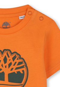 T-shirt in cotone arancione con logo ad albero verde scuro, scollatura rotonda e due bottoni a pressione sulla spalla per una facile vestibilità.