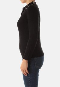 Maglione nero a maniche lunghe con un design a maglia testurizzata, dotato di un colletto borchiato. Indossato con jeans aderenti blu, vista laterale.