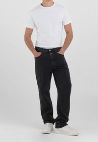 Schwarze Jeans mit gerader Beinform und Fünf-Taschen-Design, kombiniert mit einem taillierten weißen T-Shirt und weißen Sneakern. Einfarbiger Hintergrund.