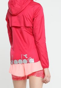 Bright pink lightweight jacket with a hood, featuring a flap at the back and silver logo at the hem. Paired with patterned pink shorts.