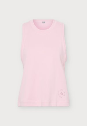 Une femme se tient face à l’objectif, vêtue d’un haut rose sans manches, d’un pantalon de jogging gris foncé et de baskets blanches, sur un fond uni clair.