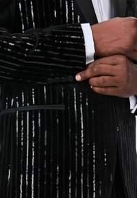 Black velvet blazer with vertical silver sequins, slim fit. Phantom pocket and subtle button detailing on the cuff, white shirt cuff visible.