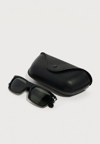 Μαύρα ορθογώνια γυαλιά ηλίου με σκούρους φακούς δίπλα σε μια μαύρη δερμάτινη θήκη με κούμπωμα, σε απλό λευκό φόντο.