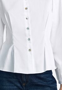 Balta pogājama krekls ar krokojumu un sešām pasteļtoņu pogām priekšpusē. Audums šķiet gluds ar strukturētu piegriezumu.