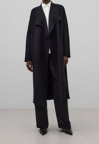 Pitkä musta takki pehmeästä kankaasta, jossa on laskostettu avoin etuosa, leveät hihat ja rento siluetti, yhdistetty mustiin housuihin ja kenkiin.