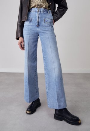 Personne portant un jean large taille haute bleu clair, une veste en cuir noire, une chemise fleurie et des bottes noires avec embouts dorés, debout sur un sol gris.