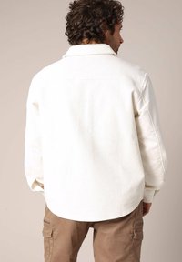 Chemise en velours côtelé blanche avec un col pointu et un ourlet arrondi, portée sur un pantalon cargo marron, mettant en valeur une texture à côtes verticales.