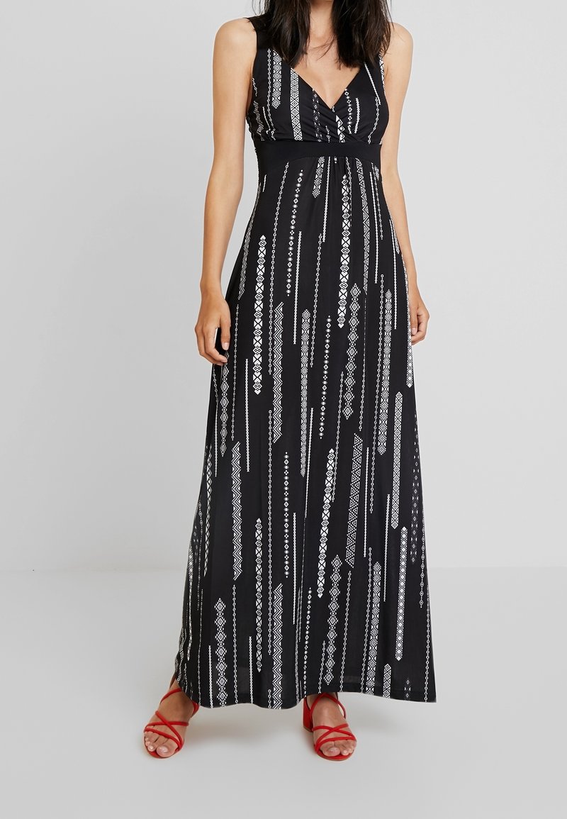Femme portant une robe longue noire sans manches avec des motifs géométriques blancs verticaux et des sandales rouges à brides, debout sur un fond gris.
