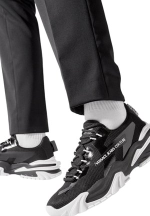 Personne portant des baskets chunky noires et blanches Versace Jeans Couture avec des chaussettes blanches et un pantalon noir, en train de marcher sur un fond blanc.