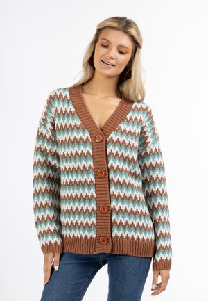 Donna che indossa un cardigan lavorato a maglia con motivo a zigzag marrone, bianco e teal con quattro bottoni, abbinato a jeans blu, in piedi davanti a uno sfondo bianco.
