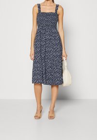 Navyblauwe bloemenjurk met vierkante halslijn, gerimpelde taille en schouderbandjes, die tot net onder de knie valt. Gecombineerd met beige muiltjes en tas.