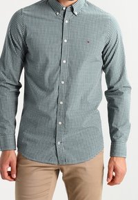 Man in een goed passende groen en witte geruite overhemd met lange mouwen, gecombineerd met beige broek, staand tegen een effen achtergrond.