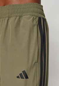 Olivenfargede treningsshorts med elastisk livlinje, med svarte sidestreker og en svart logo trykt på det nedre beinet.