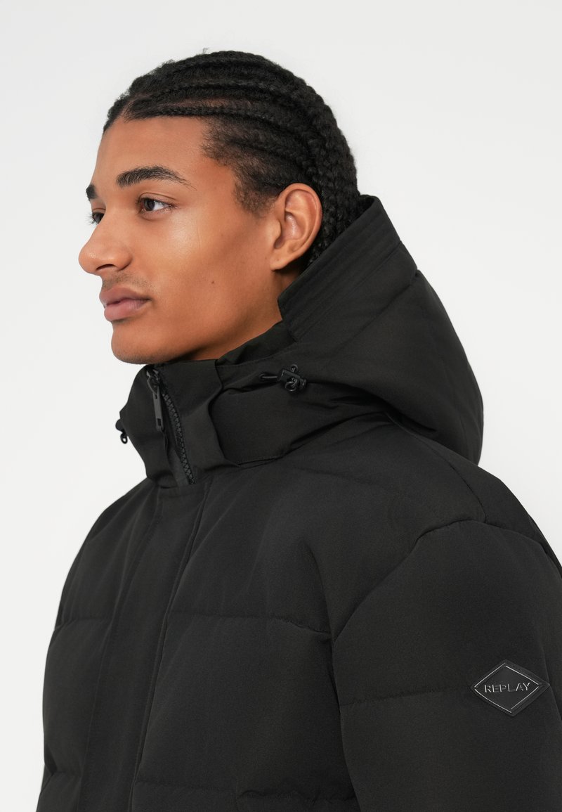 Schwarze Daunenjacke mit hohem Kragen, Reißverschluss und verstellbarer Kapuze. Verfügt über eine gepolsterte Textur und ein Logo-Patch am Ärmel.