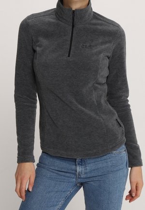 Femme portant un pull en polaire gris foncé avec une fermeture éclair frontale et un jean bleu clair, debout devant un fond uni.