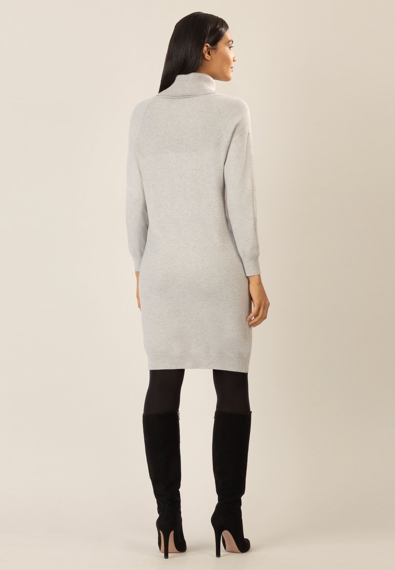 Frau in einem hellgrauen langärmeligen Rollkragenkleid, mit schwarzen Strumpfhosen und schwarzen hochhackigen Kniebots, die sich von hinten präsentiert.