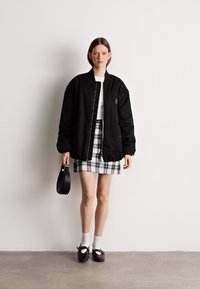 Μαύρο μπουφάν bomber με φερμουάρ μπροστά, συνδυασμένο με καρό μίνι φούστα. Το μοντέλο φοράει λευκές κάλτσες και μαύρες γόβες, κρατώντας μια μικρή τσάντα.