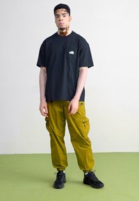 T-shirt preto de mangas curtas com bolso; calças cargo verde-azeitona com múltiplos bolsos; ténis pretos; estilo minimalista sobre um fundo neutro.