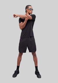 T-shirt de sport noir et shorts cargo, associés à des baskets noires et des lunettes de soleil. L'arrière-plan est gris, le sujet étire les bras.