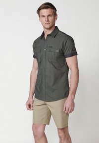 Camisa de manga corta de color verde oliva, hecha de una tela ligera, con dos bolsillos en el pecho, cuello abotonado y acentos en azul marino contrastantes.