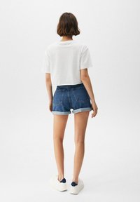 Camiseta blanca cortada, shorts de mezclilla con dobladillo enrollado y zapatillas blancas con acentos en navy. Conjunto simple y casual visto desde atrás.