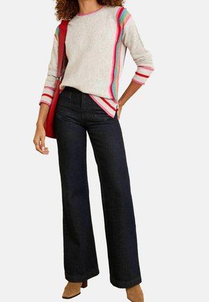 Kobieta ubrana w jasny sweter w kolorowe paski, czarne szerokie spodnie jeansowe, beżowe buty i niosąca czerwoną torbę na ramię.