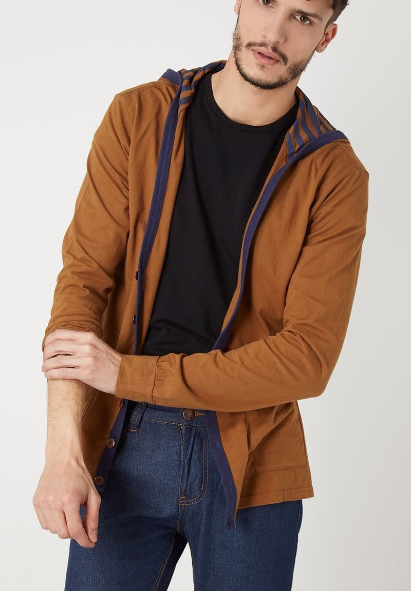 Chaqueta marrón con cremallera y capucha a rayas azul marino, hecha de algodón. Presenta un ajuste relajado con cierre de botón y mangas largas. Combinada con jeans oscuros.